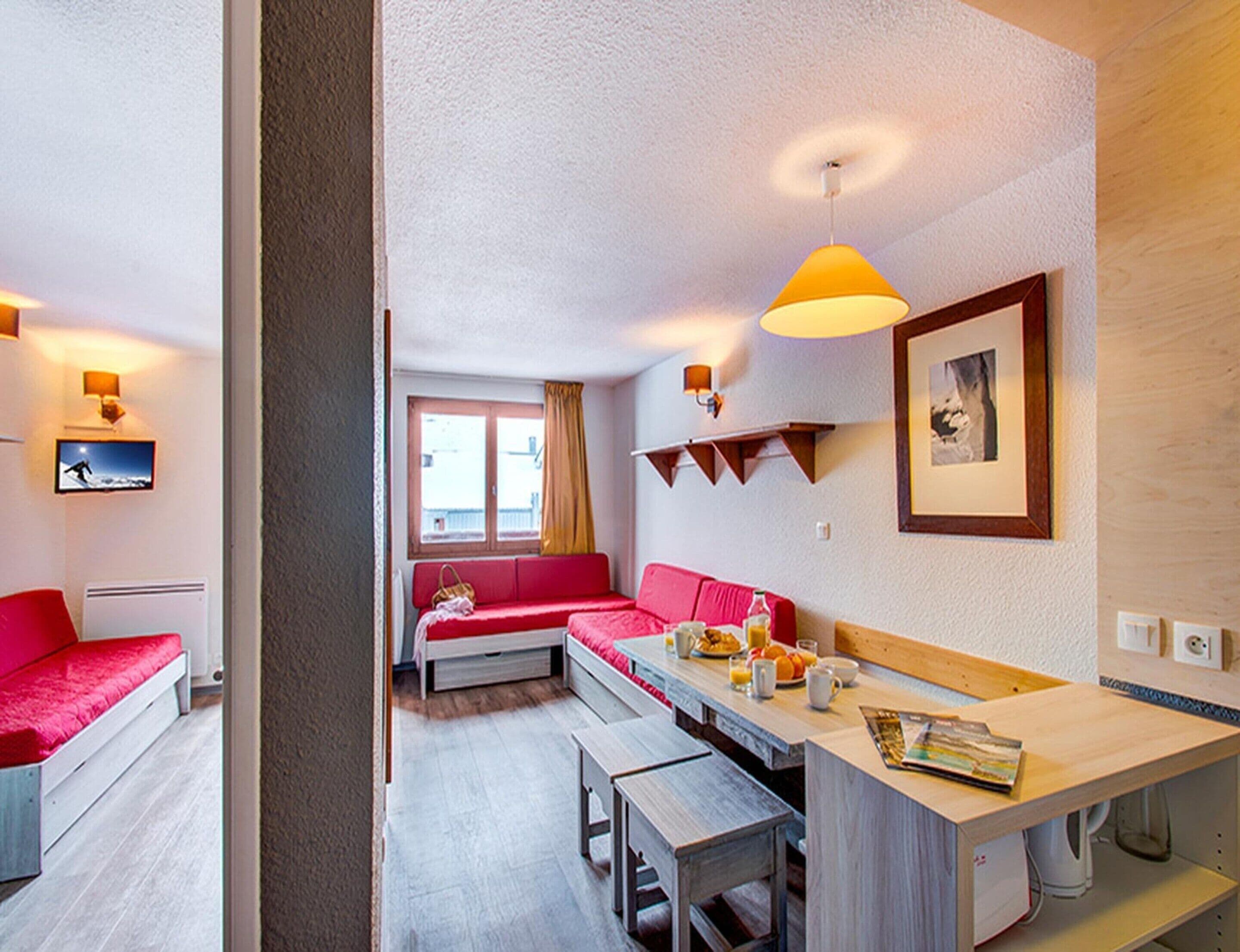 A compact hotel room with a kitchenette, a red sofa, a dining table, and a TV on the wall.