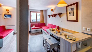 Habitación de hotel compacta con kitchenette, un sofá rojo, una mesa de comedor y un televisor en la pared.