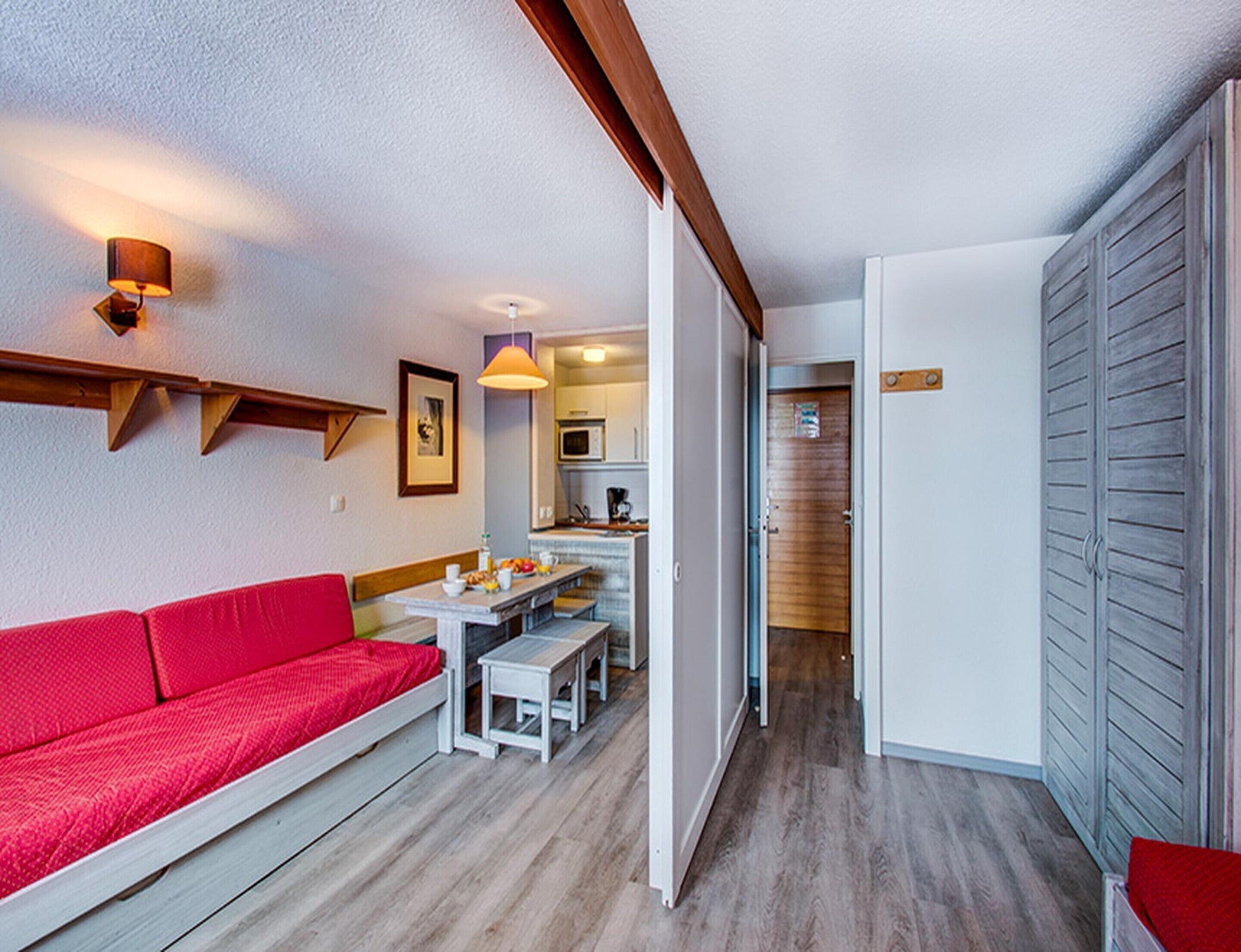 A compact living space with a red sofa, a dining table with benches, a kitchenette, and a wardrobe.