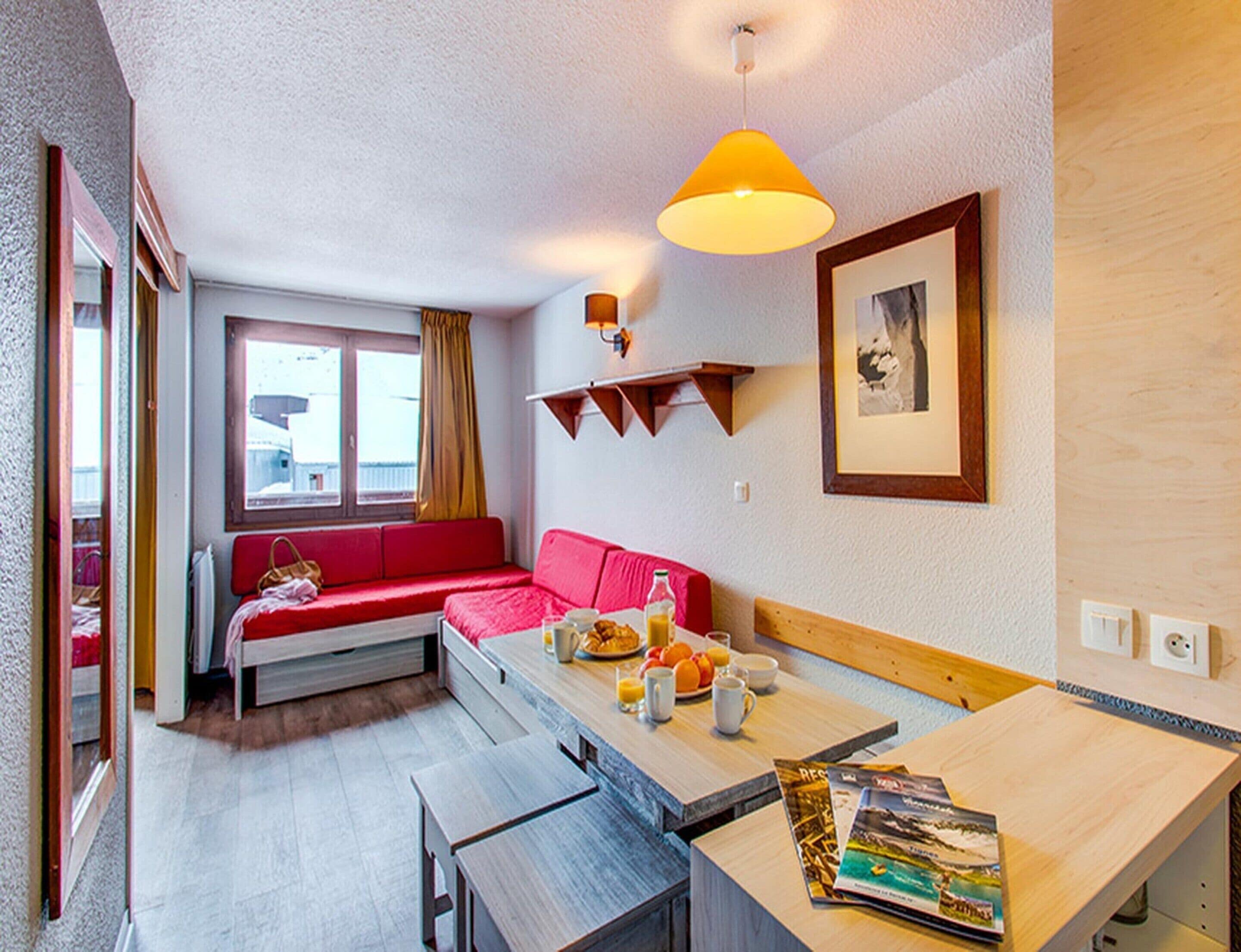 A compact room with a red sofa, a wooden dining table, a hanging lamp, and a framed picture on the wall.