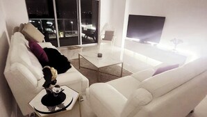 A modern living room with a white sofa, a coffee table, and a large TV screen. - Waterfront apartment (Fort Myers)