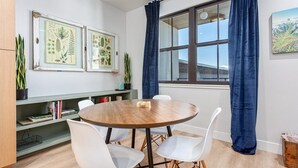 Ein Zimmer mit einem runden Holztisch, weißen Stühlen, einem Bücherregal, gerahmten Pflanzenbildern und einem Fenster mit Vorhängen.