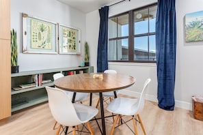 Una habitación con una mesa redonda de madera, sillas blancas, una estantería, grabados botánicos enmarcados y una ventana con cortinas.