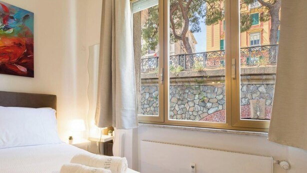 Quarto de hotel com cama, mesa de cabeceira, luminária e janela com vista para uma parede de pedra e árvores.