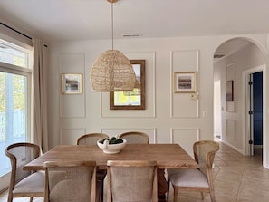 Un coin repas avec une table en bois, des chaises en osier, un luminaire suspendu en fibres tressées et des tableaux encadrés accrochés au mur.