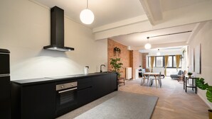 A modern kitchen with a black island, a brick accent wall, and a dining area with a table and chairs. - Grand Central Station 2br/14p Apartment (Antwerp)