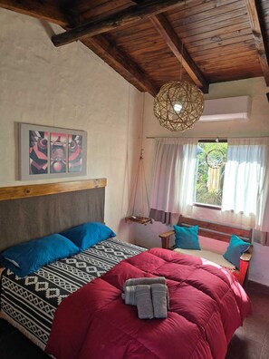 A bedroom with a wooden ceiling, a bed with patterned bedding, a hanging light fixture, and a window with curtains. - Portal de la Ovejería (Tafi del Valle)
