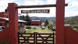 A rustic gate with a sign reading 'PORTAL de la OVEJERIA' and a view of a building with a red roof and parked cars in the background. - Portal de la Ovejería (Tafi del Valle)