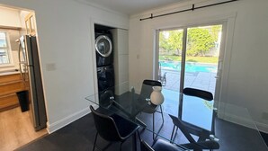 Ett modernt kök med inbyggd tvättmaskin och torktumlare, en matplats med ett glasbord och stolar samt en skjutdörr i glas som leder ut till en trädgård med pool.
