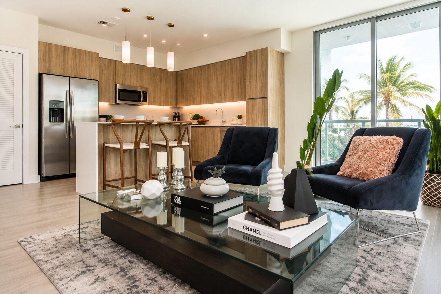 A modern living room with a glass coffee table, a dark armchair, and a view of the kitchen and outdoors.