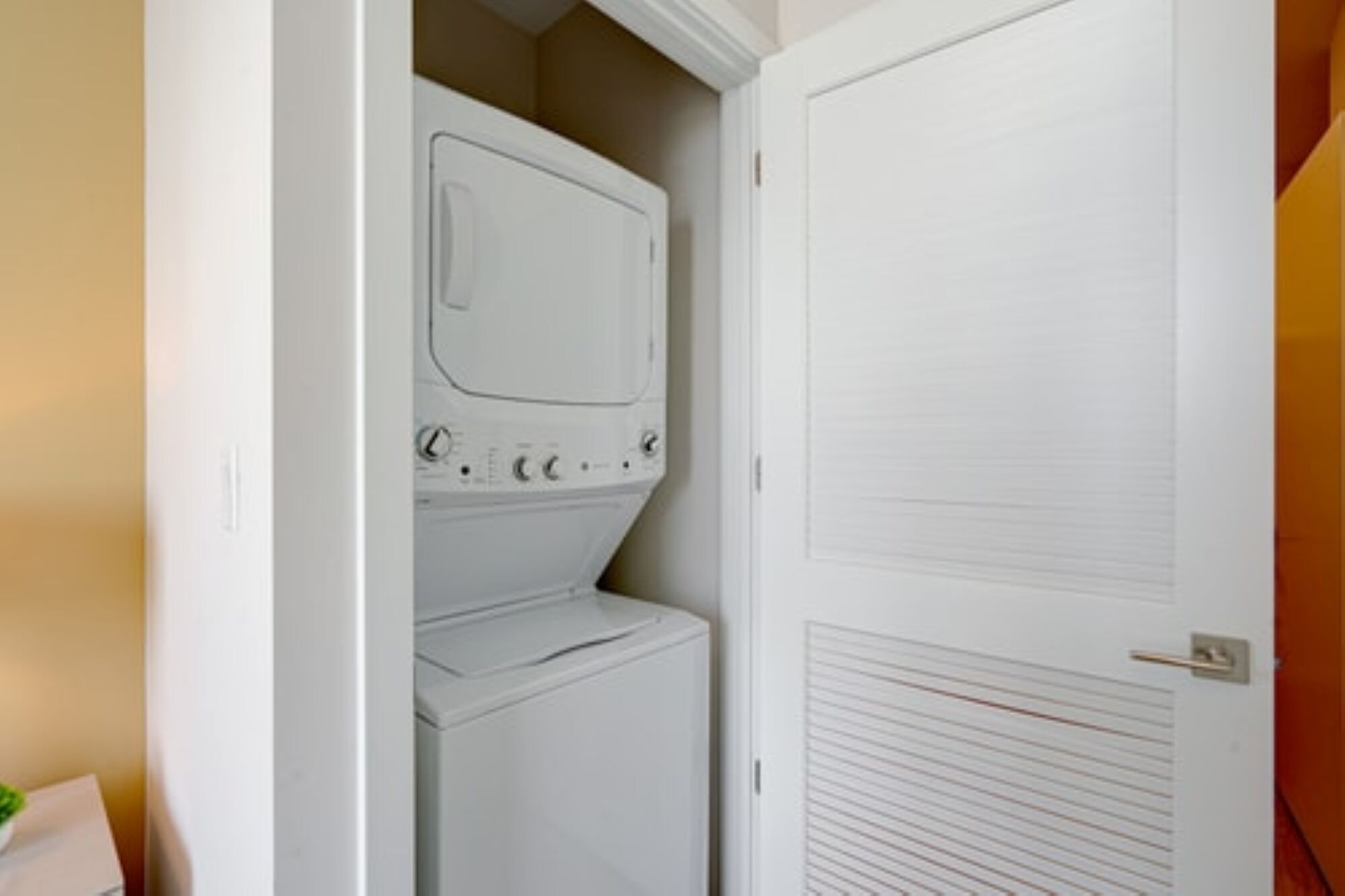Uma lavanderia com máquina de lavar e secadora empilhadas, uma porta com ventilação e uma vista parcial de uma parede.