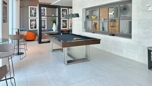 Uma mesa de bilhar moderna em ambiente interno, com taco, área de bar com bancos e parede com quadros e prateleiras.