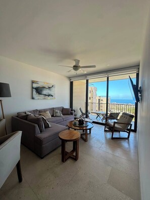 Ein modernes Wohnzimmer mit einer Couch, einem Couchtisch und Sesseln, einem Deckenventilator und einem großen Fenster mit Blick auf Gebäude und das Meer.