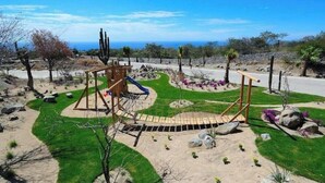 Ein Spielplatz mit Rutsche und Klettergerüsten, umgeben von begrünten Flächen mit Steinen und Kakteen.