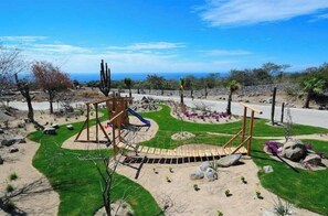 Ein Spielplatz mit Rutsche und Klettergerüsten, umgeben von begrünten Flächen mit Steinen und Kakteen.