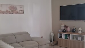 Un saló amb un sofà seccional beix, un televisor de pantalla plana muntat a la paret, una taula auxiliar petita i una estanteria amb diversos objectes decoratius.
