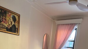 A bedroom with a bed, pillows, a painting, a ceiling fan, a window with curtains, and a vase with flowers. - Delightful 1-bedroom suit in boutique guesthouse with AC in vibrant New York (New York)