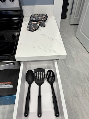 Un plan de travail de cuisine surmonté d’un plateau en marbre blanc, avec une cuisinière et un tiroir contenant des ustensiles de cuisine noirs.