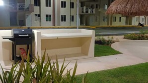 A well-lit outdoor area with a barbecue grill, a thatched-roof structure, and a multi-story building in the background. - APARTMENT IN CANCUN WITH SWIMMING POOL IN A CONDOMINIUM (Cancún)