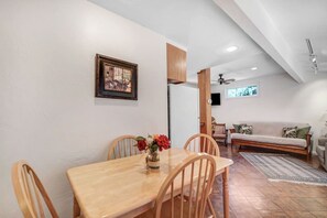 A dining area with a wooden table and chairs, a vase with flowers, a painting on the wall, and a living area with a sofa and a TV. - Arches - Olive Grove Cottage (Sonoma)