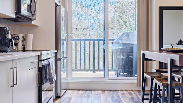 A modern kitchen with a large window, a dining area with stools, and a view of the outdoors. - k211- AC, Pool/Hot Tub, Parking, BBQ, View (Canmore)