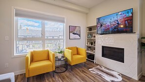 A living room with a fireplace, a large TV, two yellow armchairs, a small round table, and a view of a cityscape through large windows. - k211- AC, Pool/Hot Tub, Parking, BBQ, View (Canmore)