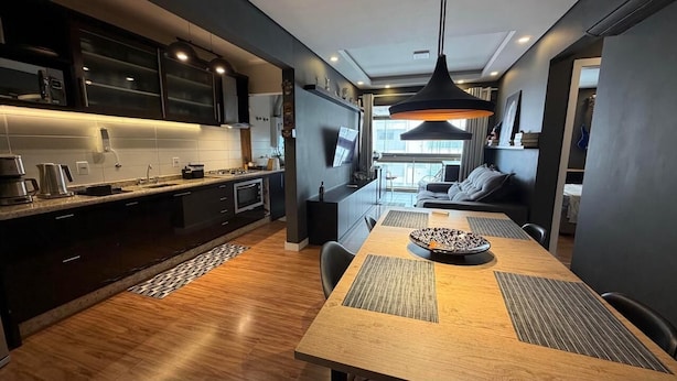 Une cuisine moderne avec des meubles de rangement sombres, une table à manger en bois et un luminaire suspendu.