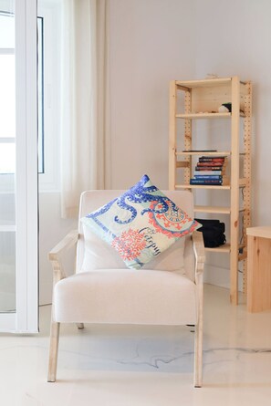 A room with a white armchair, a patterned throw pillow, a wooden bookshelf, and a small table. - Caicos Calm: Serene 2BR Condo in Whitby, North Caicos - a Garden Isle Getaway (Whitby)