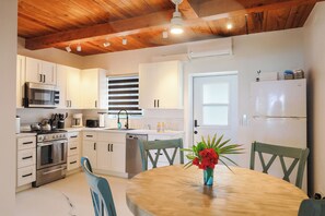 A kitchen with white cabinets, a microwave, stove, and a sink. There is a dining table with chairs and a vase of flowers. - Caicos Calm: Serene 2BR Condo in Whitby, North Caicos - a Garden Isle Getaway (Whitby)