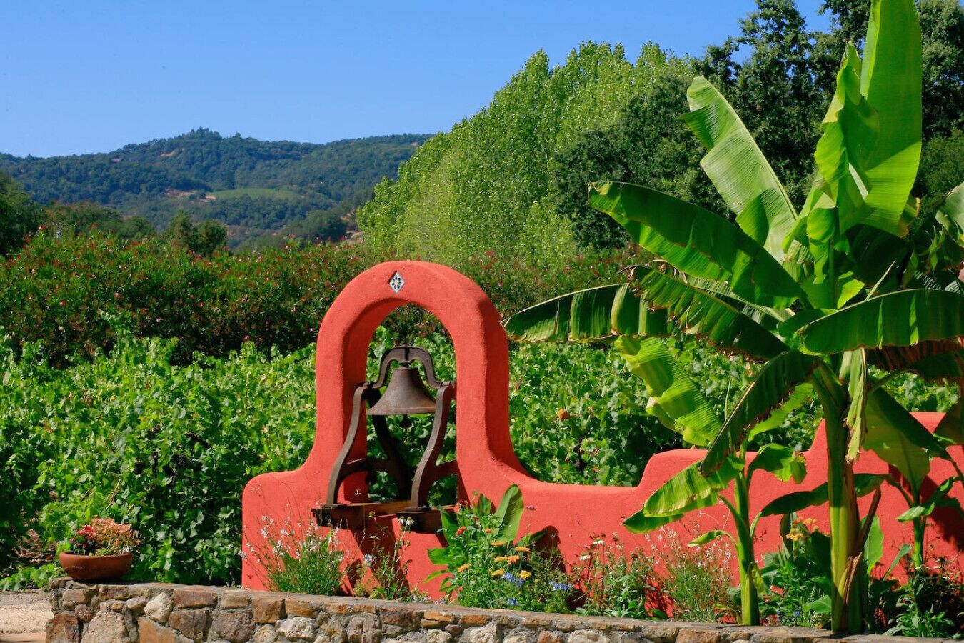Un campanario con un arco rojo, rodeado de abundante vegetación y un muro de piedra.