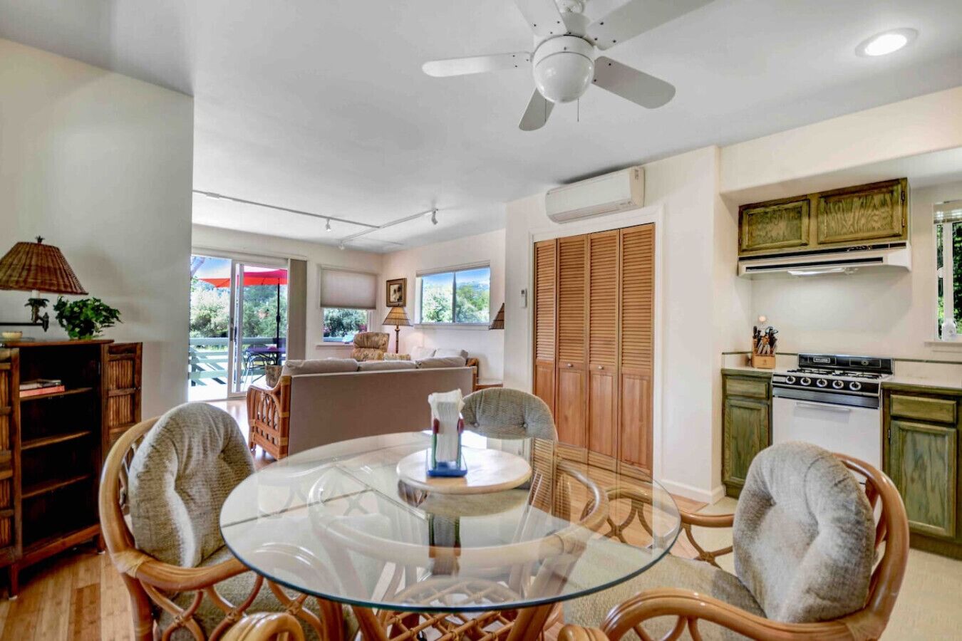 A dining area with a glass table, wicker chairs, a ceiling fan, and a kitchen with wooden cabinets and a stove.