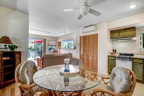 Un coin repas avec une table en verre, des chaises en osier, un ventilateur de plafond et une cuisine dotée de meubles en bois et d’une cuisinière.