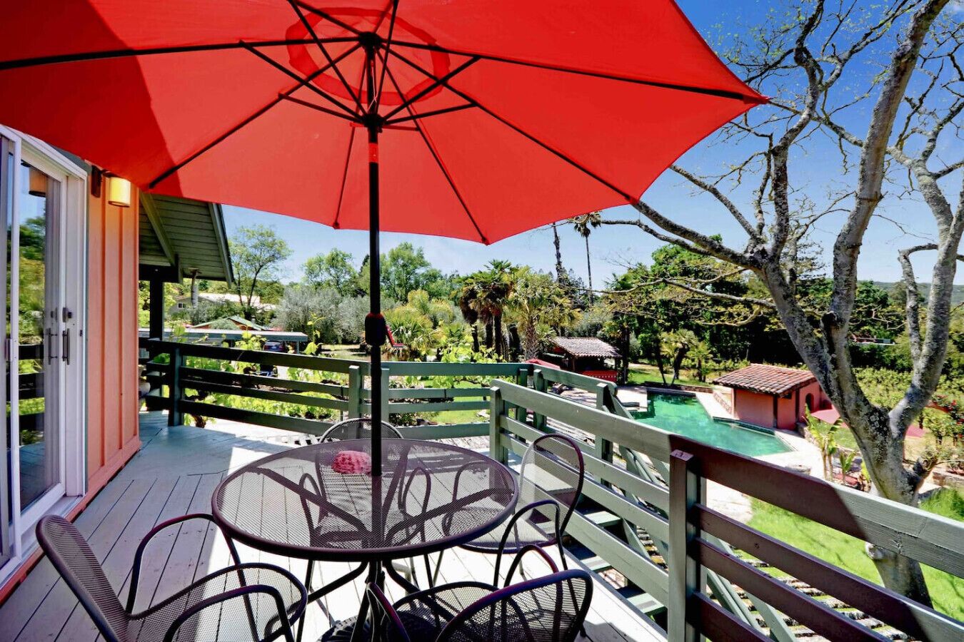 A covered outdoor area with a table and chairs, a red umbrella, and a view of a pool and surrounding greenery.