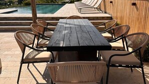 Une terrasse extérieure avec une table en bois et des chaises en osier, donnant sur une piscine et sur des collines pittoresques.