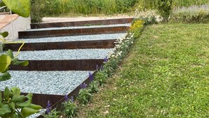 Un sentiero con gradini che conduce attraverso un giardino con diverse piante e un campo sullo sfondo.