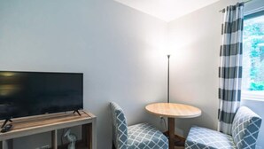 Ein Eckzimmer mit Flachbildfernseher auf Holzständer, zwei gemusterte Sessel, ein kleiner runder Tisch und ein Fenster mit gestreifter Vorhang.