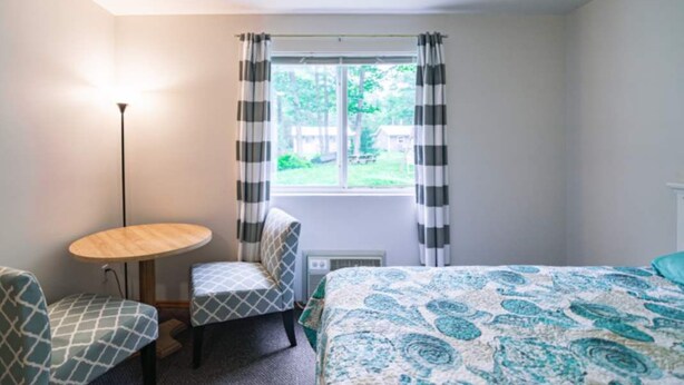 Ein Schlafzimmer mit einem Bett, einem Sessel, einem kleinen Tisch und einem Fenster mit Vorhängen.