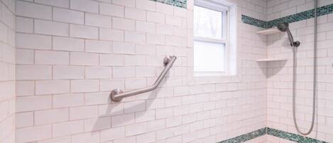 Een badkamer met witte betegeling in metro-stijl, groene mozaïekranden, een raam, een douche en een handgreep.