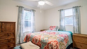 A bedroom with a bed, a wooden dresser, a nightstand, a ceiling fan, and two windows with curtains. - Cabin #7 Wishing Star (Iosco County)