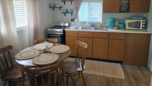 A compact kitchen with wooden cabinets, a stove, a microwave, and a dining table with chairs. - Cabin #2 Water's Edge (Iosco County)