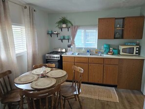 A compact kitchen with wooden cabinets, a stove, a microwave, and a dining table with chairs. - Cabin #2 Water's Edge (Iosco County)