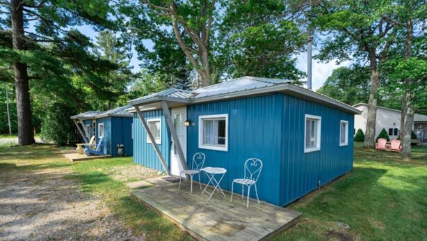 A small blue cabin with a porch, two white chairs, and a wooden deck. - Cabin #2 Water's Edge (Iosco County)