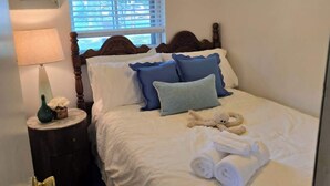 A neatly made bed with pillows and a teddy bear, a lamp on a bedside table, and a window with blinds. - Cabin #2 Water's Edge (Iosco County)