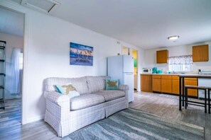 Una sala de estar con un sofá, una zona de cocina con armarios de madera, un refrigerador y una mesa de comedor.