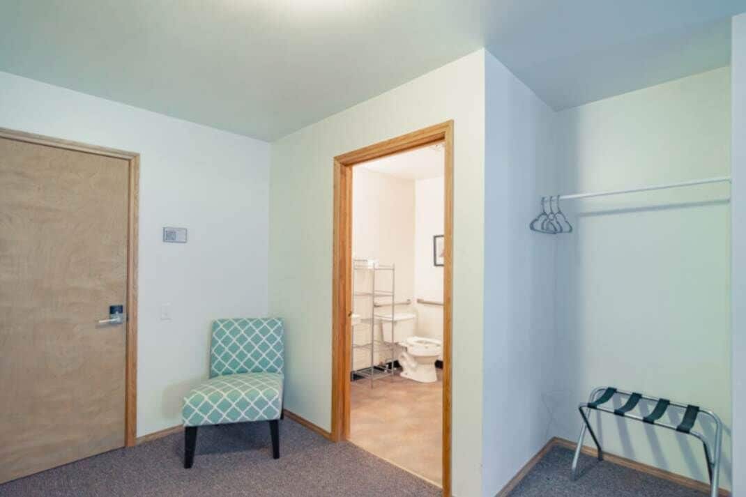 A room with a wooden door, a patterned chair, a metal rack, and a view into a bathroom with a toilet and shelves.