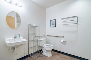 Un baño con lavamanos blanco, inodoro y espejo. Hay un estante metálico con una toalla y un cuadro en la pared.