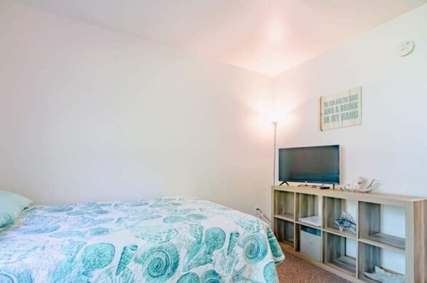 Un dormitorio con una cama, un televisor sobre un mueble, una estantería con objetos y un reloj de pared.