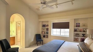 Een slaapkamer met een bed, een stoel, een plafondventilator, een raam en een boekenkast.
