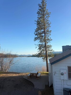 Uma cabana à beira do lago com uma mesa de piquenique, uma pinheiro alta e uma vista desobstruída do lago e montanhas distantes.