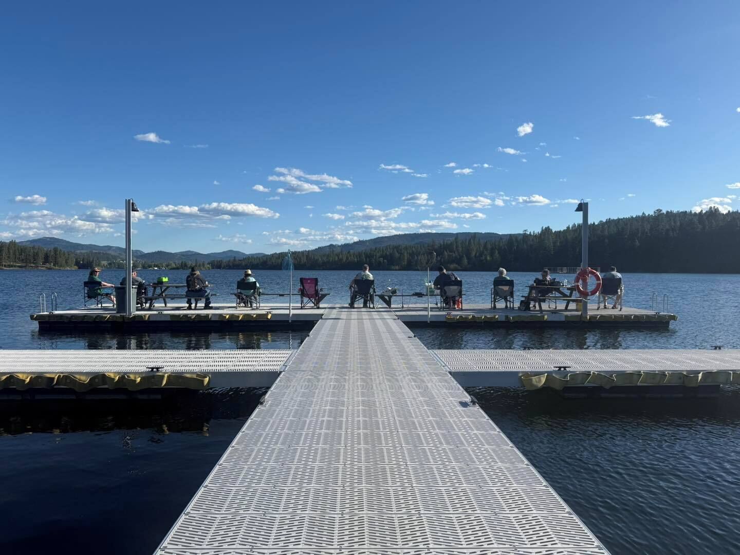 Un muelle con gente sentada, un flotador salvavidas y un lago tranquilo con montañas al fondo.